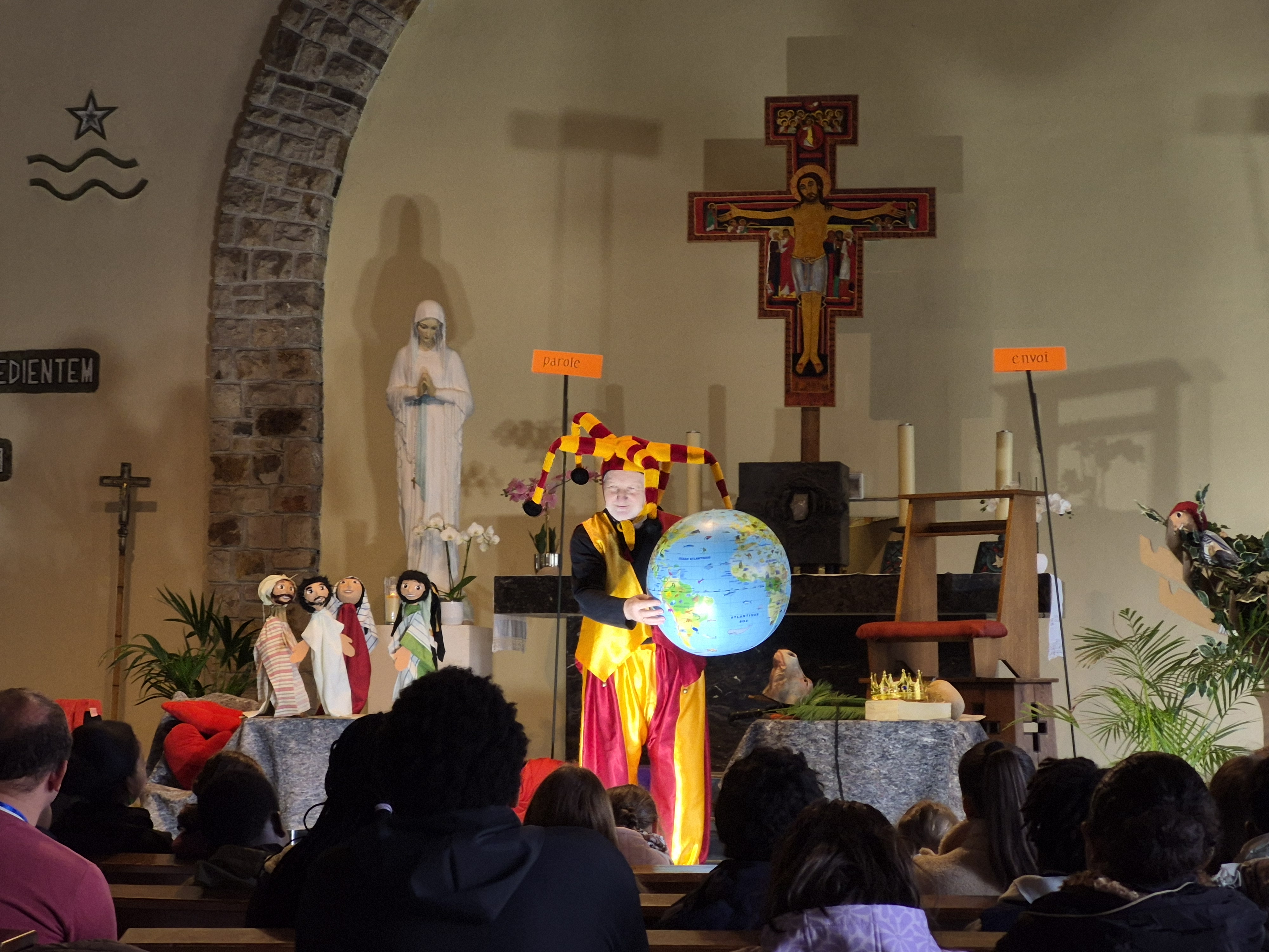 Devant l'autel, Luc Aerens, en costume de bouffon rouge et jaune, tient un globe terrestre face aux acolytes assis, vus de dos, pendant son spectacle