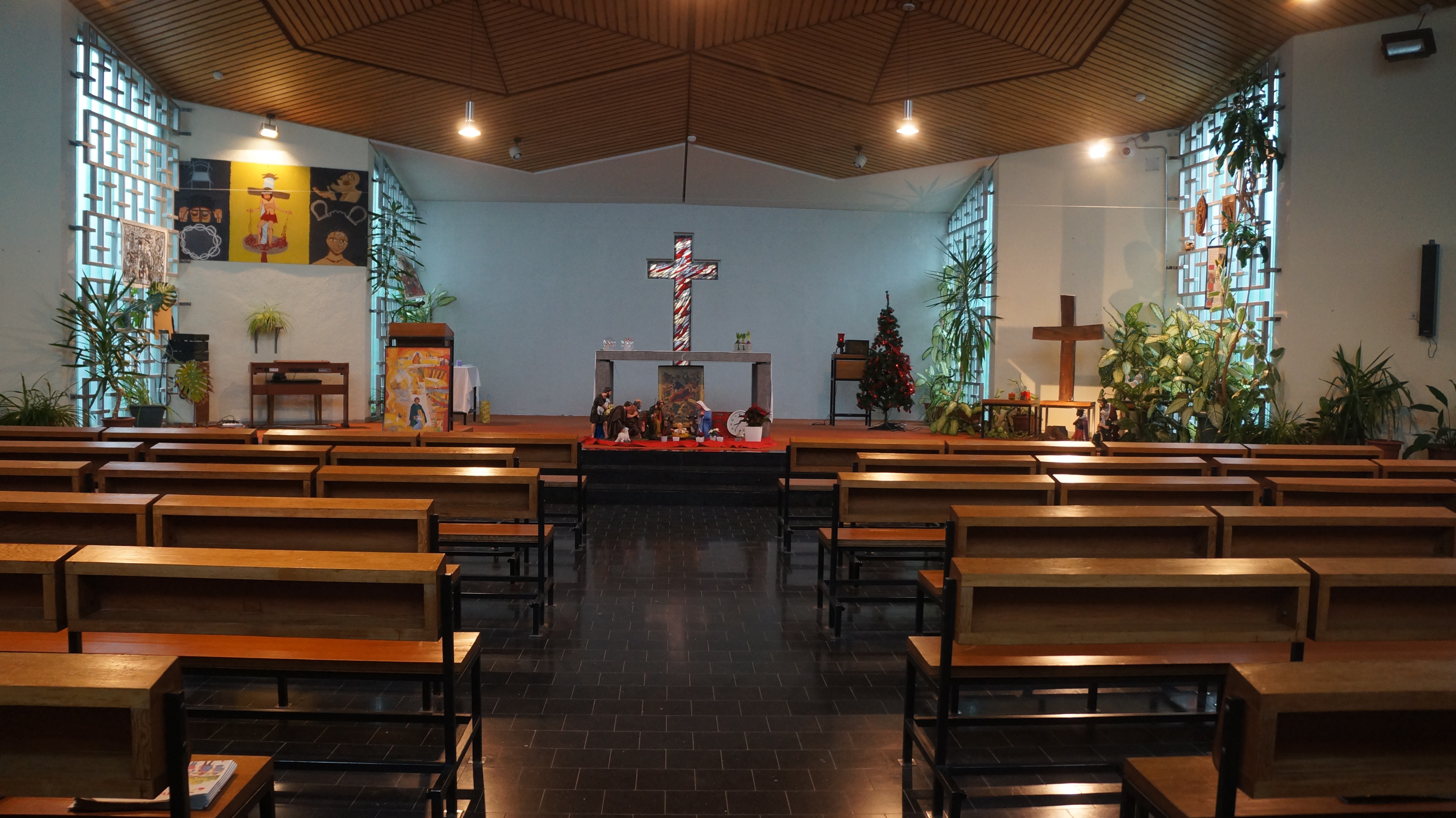 Une chapelle sous un plafond de bois, aux fenêtres de pavés de verre&nbsp;: des rangées de bancs mènent à l'autel, au pied duquel sont installés une crèche et un sapin de Noël, sous une grande croix colorée&nbsp;; des plantes vertes et des tableaux habillent les murs