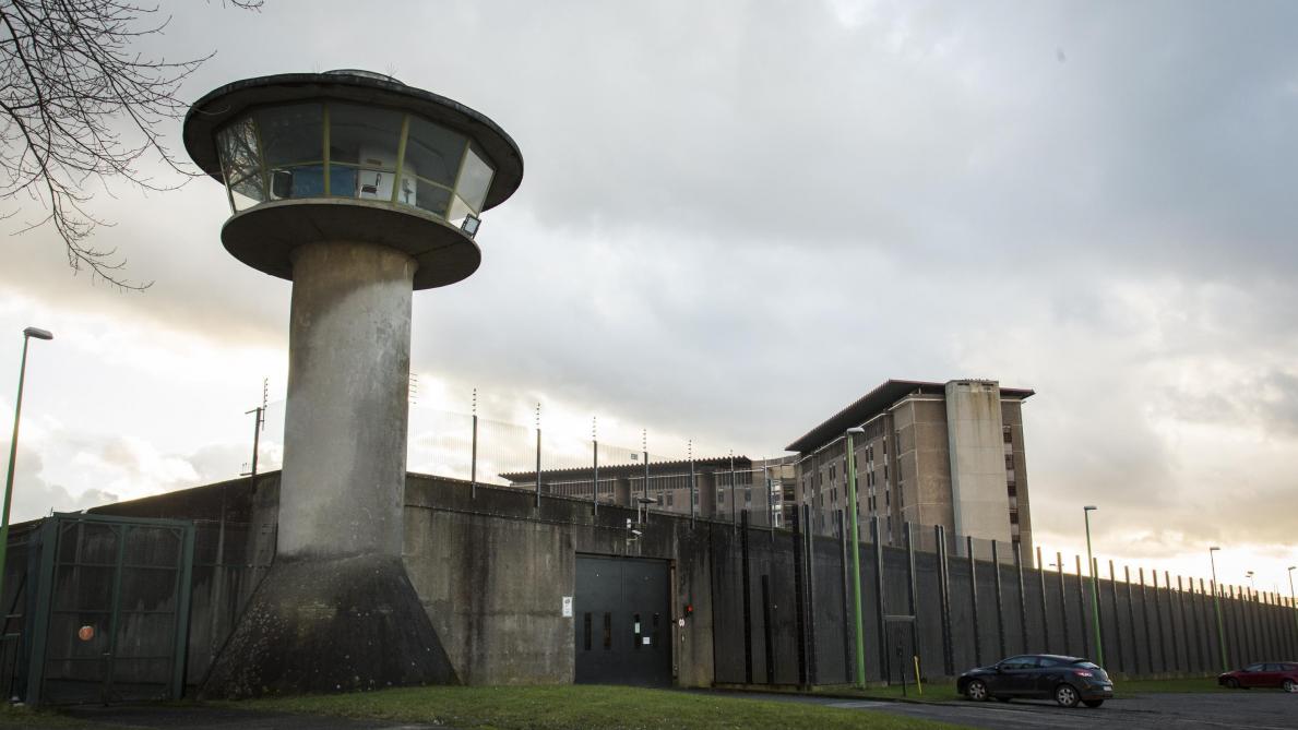 Une prison vue de l'extérieur, sous un ciel chargé&nbsp;: une tour de surveillance en béton domine un haut mur gris surmonté de grillages et de barbelés, devant de grands bâtiments de détention